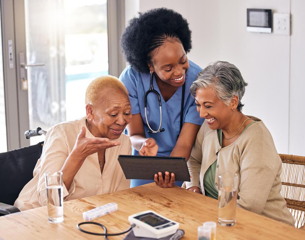 Two senior women hold a tablet and chat with a nurse while enjoying senior leaving near Lewiston, MN.